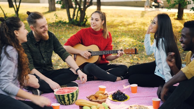 A group of young adults sitting outside on a picnic blanket, talking and playing guitar together.